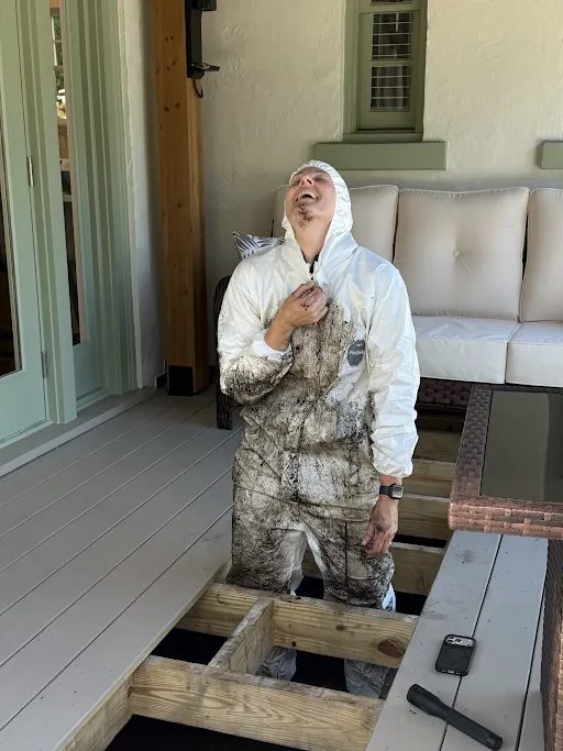 HouseScan inspector in a white protective suit laughing while climbing out from under a porch